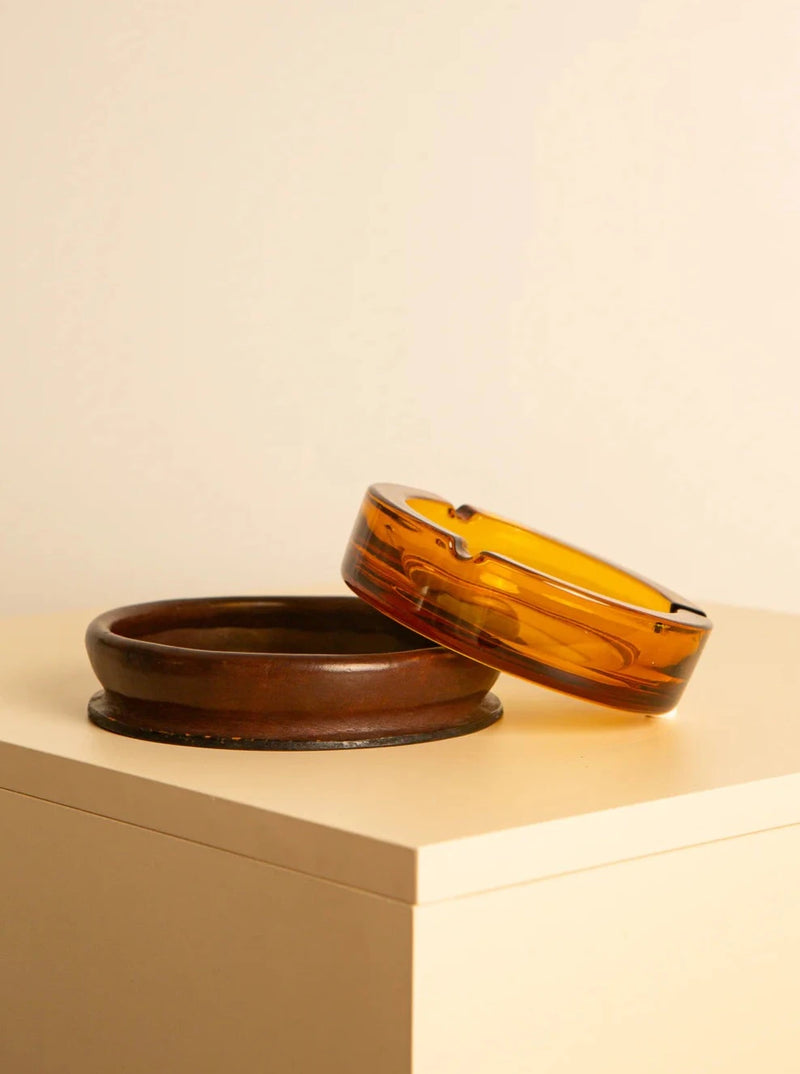 A brown Treaptyque Glass and Italian leather ashtray 60's is placed on a light-colored surface, with a yellow glass Treaptyque Glass and Italian leather ashtray 60's resting on its edge. The background is plain and softly lit, evoking retro warmth and highlighting the simplicity and contrast between the two ashtrays.
