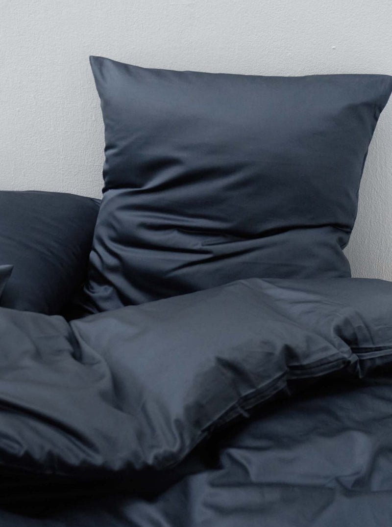 The Cotton Sateen Bed Set in Stormy by MOST, featuring dark-colored pillows and a duvet against a neutral wall, showcases a contemporary design with its smooth, matte finish—ideal for those who appreciate a refined, minimalist aesthetic.