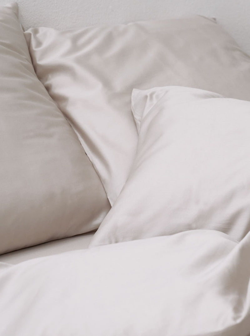 Smooth satin pillows and a MOST cotton sateen bed set in sand arranged neatly on a bed, showcasing a minimalist and comfortable bedroom setup.