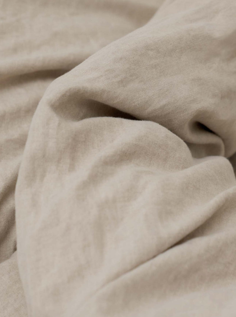 A close-up of the Washed Linen Bed Set in Natural by MOST reveals its beige, wrinkled fabric with soft texture and flowing folds. The lighting highlights subtle texture variations, enhancing its luxurious appeal for exclusive luxury interiors.
