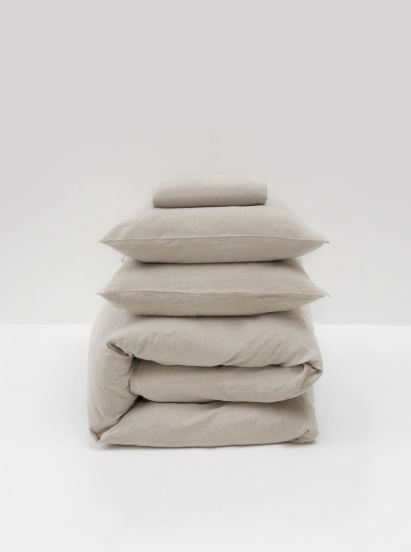 A minimalist display against a white background features the MOST Washed Linen Bed Set in Natural, showcasing neatly stacked beige bedding with two pillows and a folded duvet.