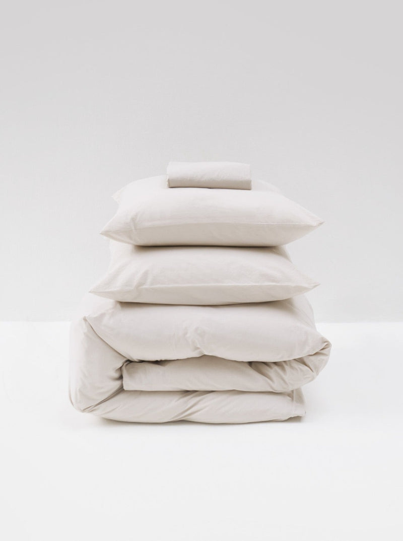 A neat stack of MOST's Washed Cotton Percale Bed Set in Sand, including two pillows, two duvets or comforter covers, and a folded towel, all in shades of white and cream, against a white background.