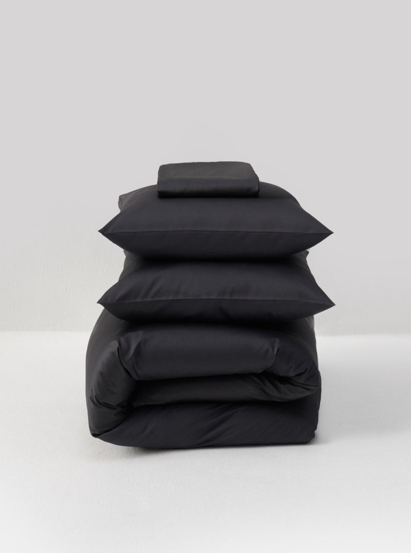 The Cotton Sateen Bed Set in Graphite by MOST is elegantly arranged on a white surface against a light gray background, featuring a folded duvet, two pillows, and a sheet. The setup exudes contemporary design and adds luxury to any bedroom.