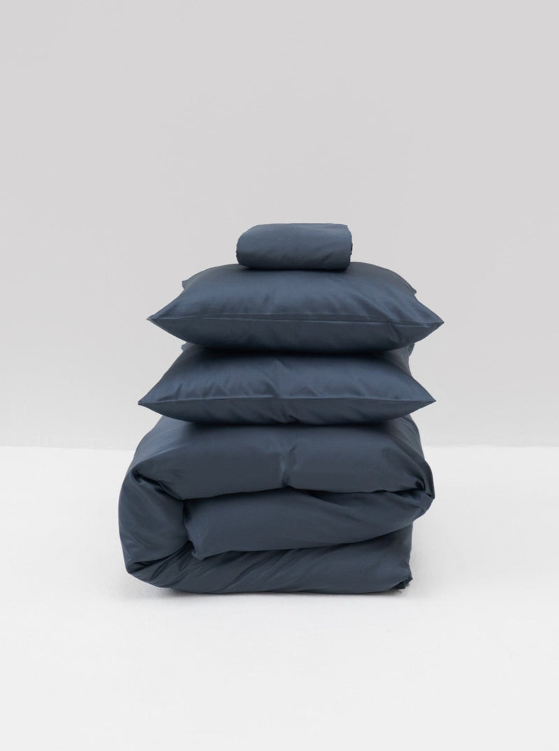 The Cotton Sateen Bed Set in Stormy by MOST, featuring a folded duvet, two pillows, and a small cushion, rests elegantly on a light gray background. This neatly arranged dark blue bedding embodies contemporary design with its luxurious touch.