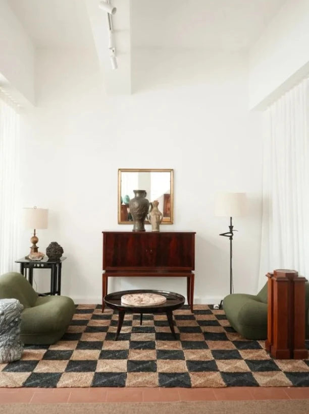 A living room with a modern aesthetic, featuring a checkered rug, two green low chairs, a dark wooden coffee table, a dark wood sideboard with vases on top, and a "Brejos" Floor Lamp by Barracuda Interiors. The walls are white with a high ceiling and minimalistic decor accented by subtle bird designs.