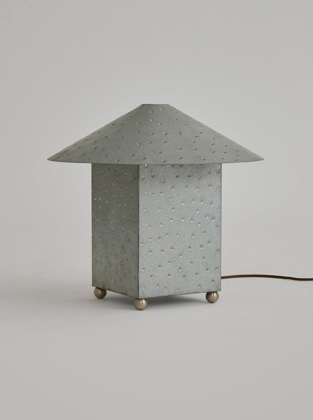 The Lin Table Lamp, Steel by MUHLY from the Lin Lighting Collection features a gray, textured, pyramid-shaped shade atop a square base. Small perforations create a starry-night effect on this hand-punched metal lamp, which is completed by four spherical feet and a brown power cord extending from the back.