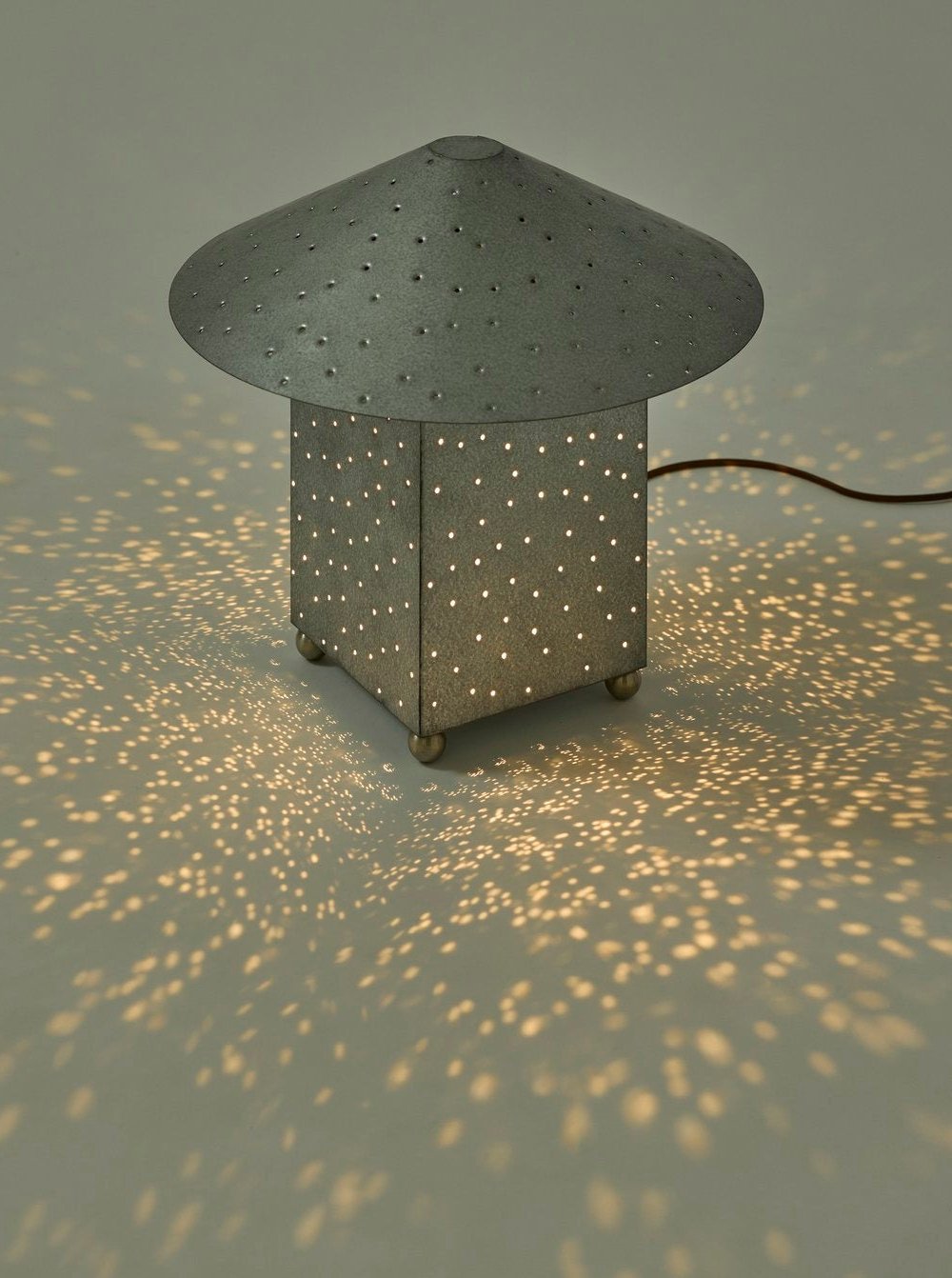 The Lin Table Lamp, Steel by MUHLY is a minimalist, square-shaped hand-punched metal lamp featuring a wide, conical top that emits a soft, warm light through numerous perforations. These holes project a starry-night effect on the surrounding surface. As part of the Lin Lighting Collection, the lamp is connected to a power cord.