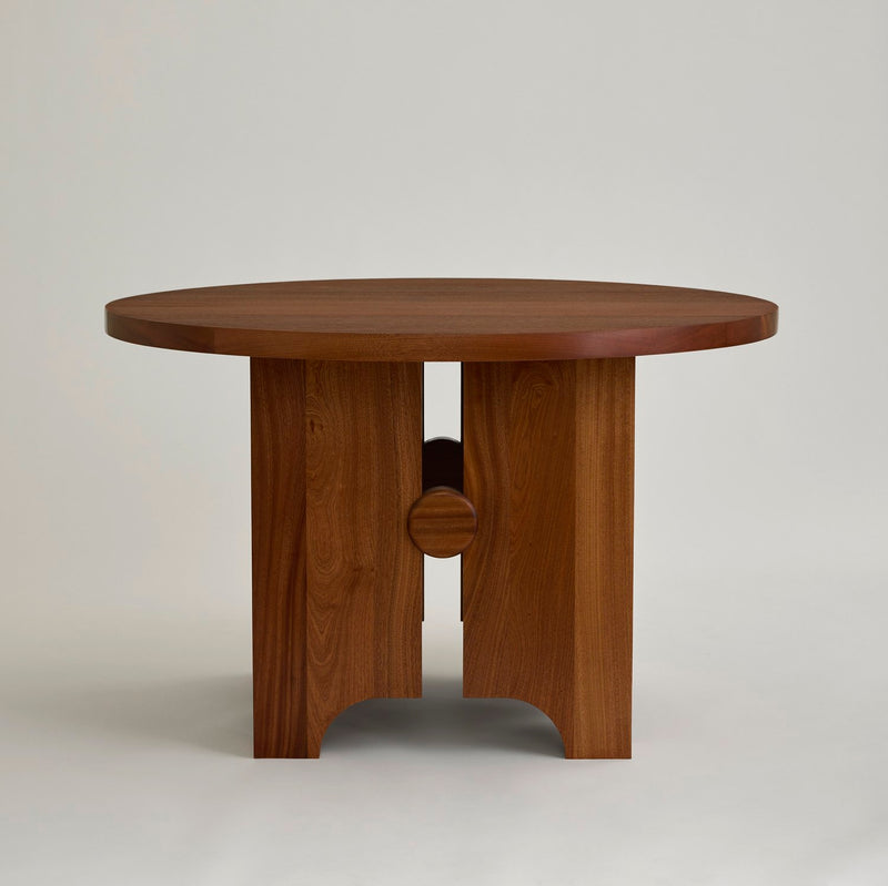 The First Light Table by MUHLY is a round wooden piece featuring a smooth top and an intricate base with four legs connected by a circular wooden support. Handmade in Philadelphia, this table boasts a natural, polished finish and stands elegantly on a neutral background.