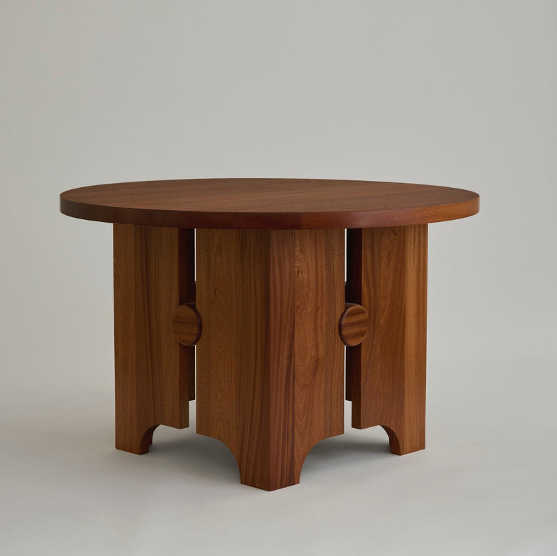 The First Light Table by MUHLY is a round wooden marvel, handcrafted in Philadelphia. It features a flat surface and an innovative base with four wide legs, each adorned with a semicircular cutout at the bottom and a rounded, decorative element halfway up. Its smooth finish makes it ideal for any independent bookstore setting.