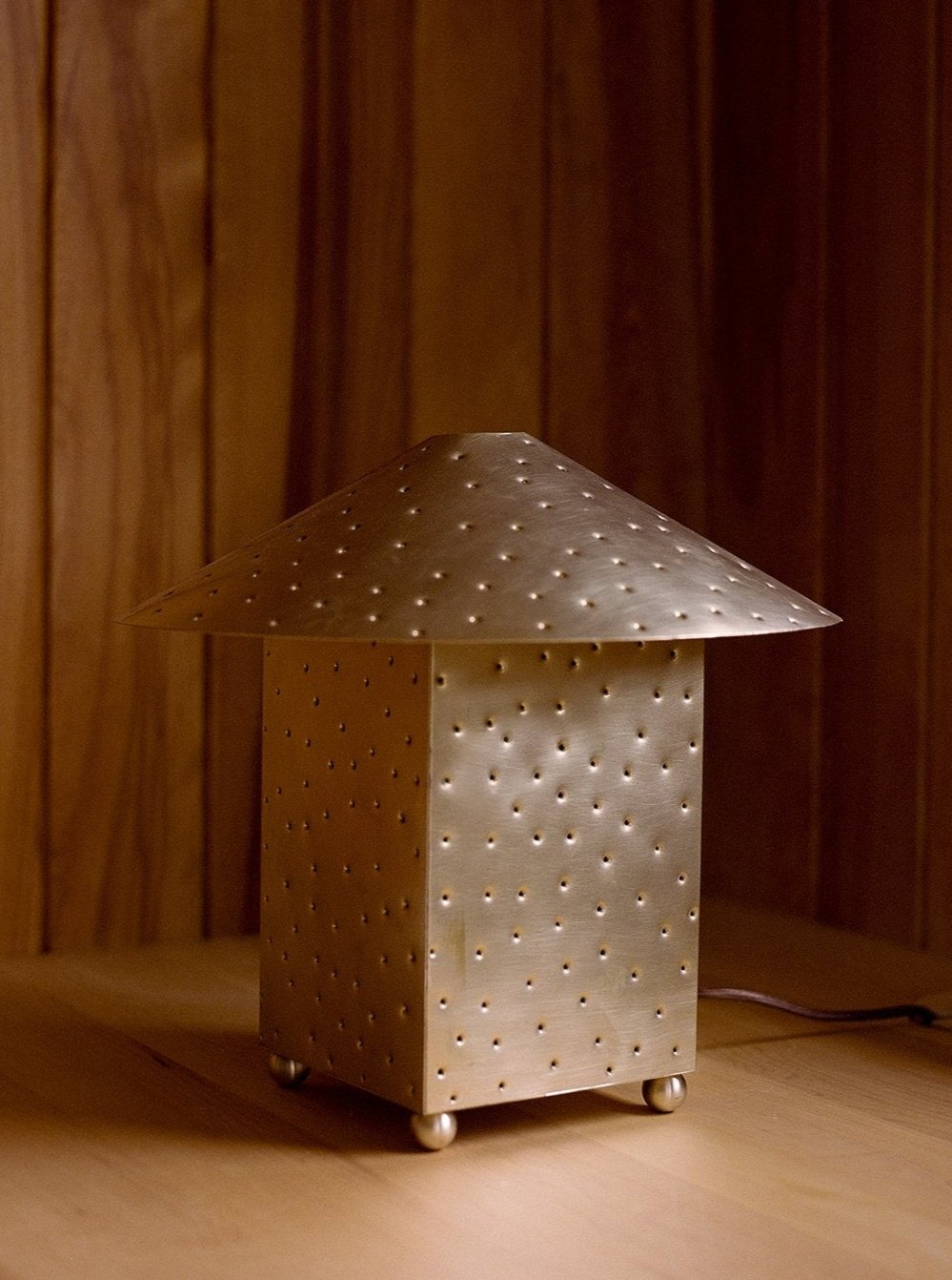 The Lin Table Lamp by MUHLY, made of steel with a perforated conical shade and rectangular base, emits a warm light. This hand-punched metal lamp features small holes that add texture and character, creating a starry-night effect as it sits on a wooden surface against a wooden backdrop.