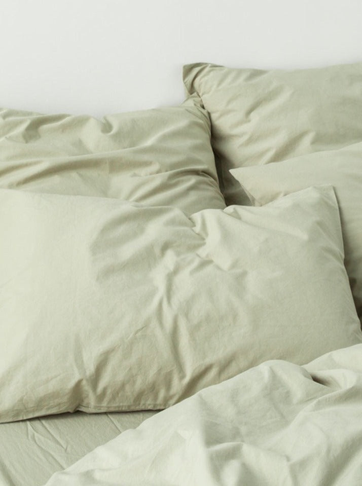 Unmade bed with rumpled MOST washed cotton percale bed set in sage and multiple pillows against a light background, conveying a cozy, relaxed atmosphere.