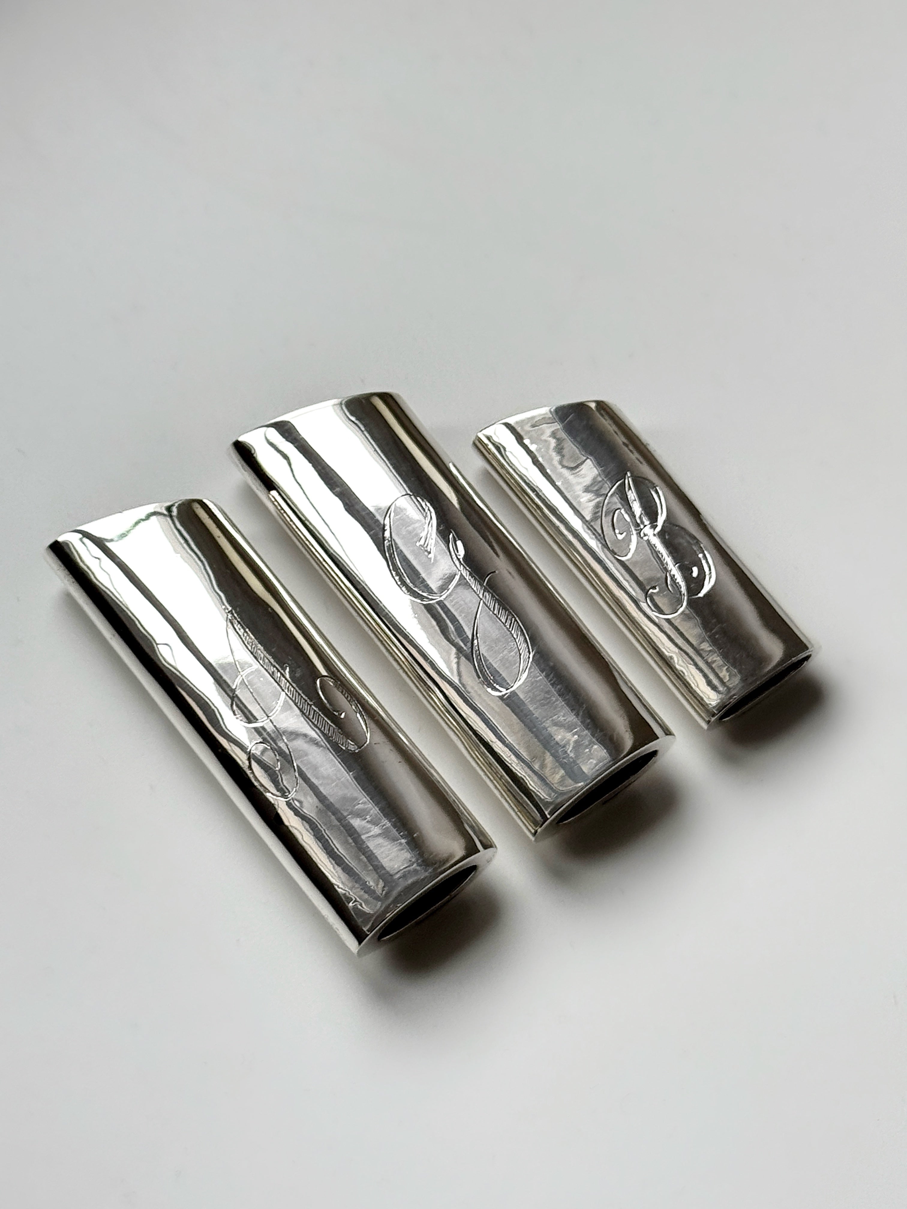 Three Pedro Ventura sterling silver lighter cases, engraved with initials "L," "E," and "B," rest on a white surface, each a bespoke heirloom in its own right.