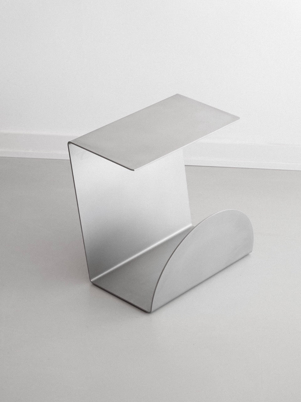 The KØGE Design Stainless Steel Side Table features a modern, minimalist look with a sleek geometric design. It has a flat top, an open curved base, and a polished surface, ideal for contemporary spaces against white walls and light floors.