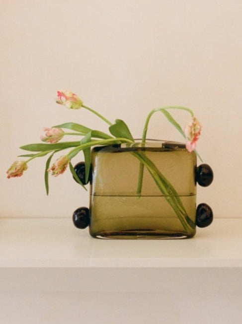 A square, translucent green Sophie Lou Jacobsen La Pierre Vase on wheels filled with water and sprouting pink tulips positioned against a neutral background.