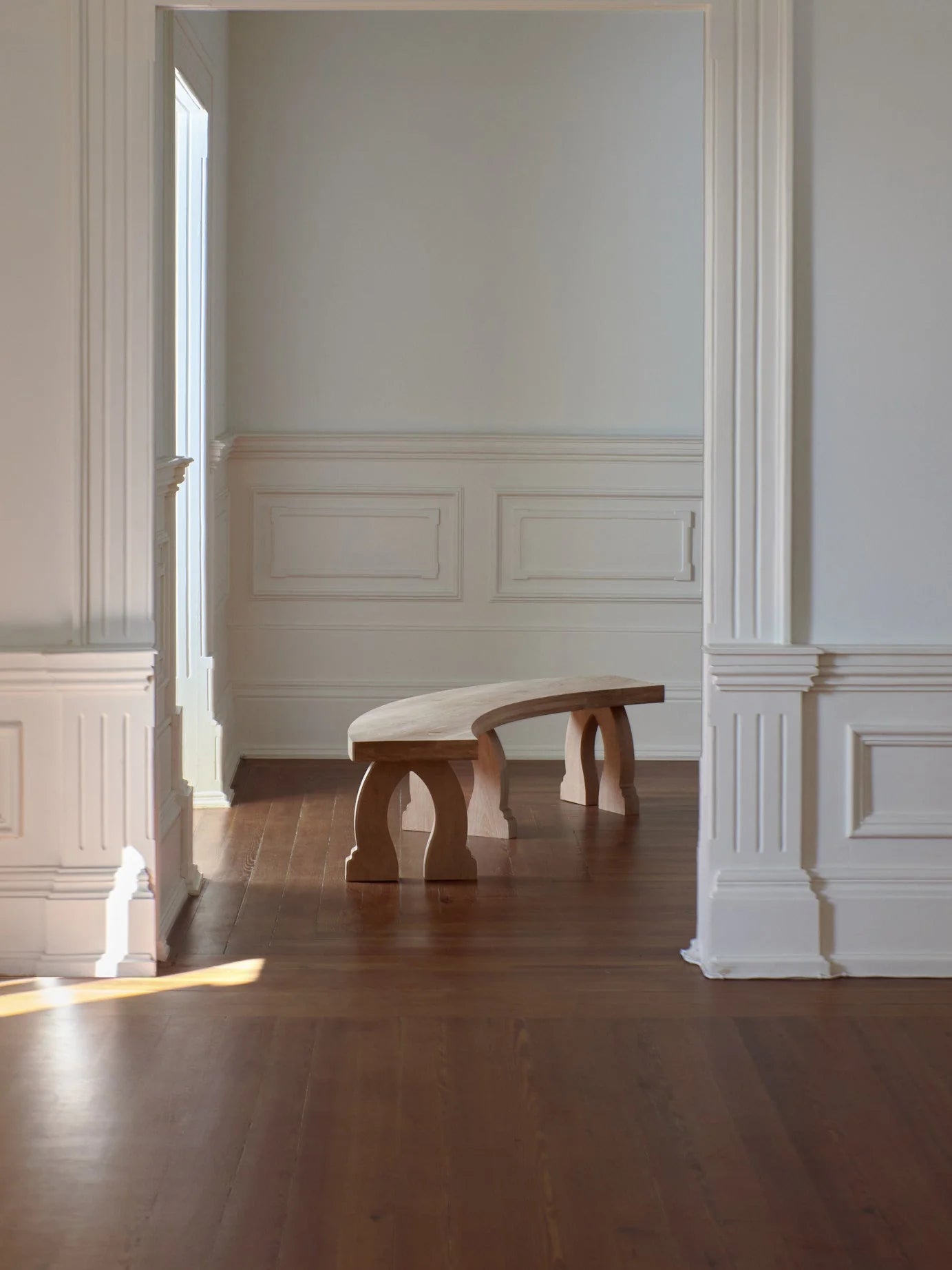 A minimalist interior features white-paneled walls, a high ceiling, and a wooden floor. At the center of the room is a Project 213A "Sintra" Curved Bench in Oak, crafted by expert artisans. Sunlight streaming through a large window enhances the calm and serene atmosphere.