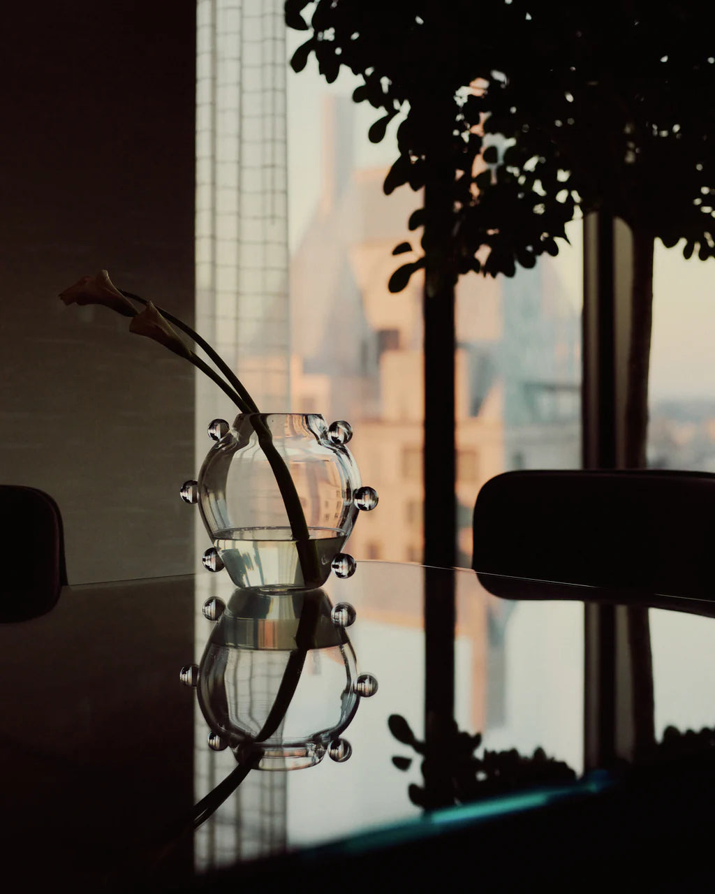 The La Lumiere Vase by Sophie Lou Jacobsen, featuring two calla lilies, rests on a reflective table with a blurred cityscape and tree glimpsed through a large window in the background.