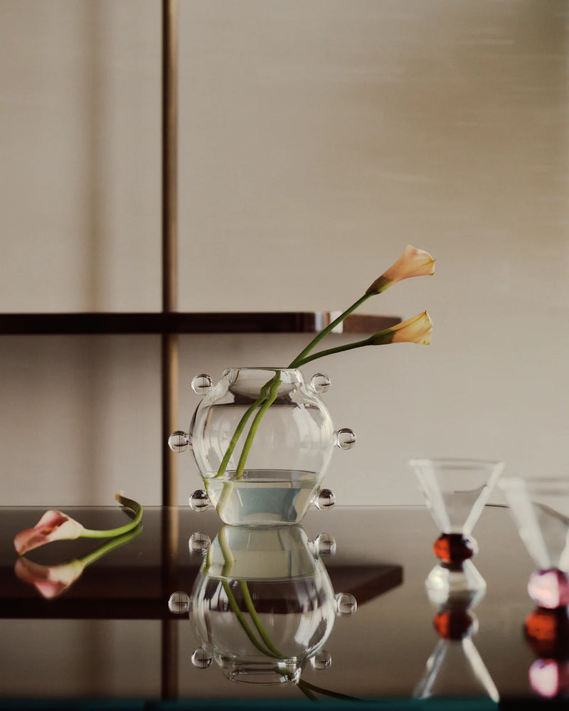 The La Lumiere Vase by Sophie Lou Jacobsen, featuring round knobs, displays two pink calla lilies on a reflective surface with one lily beside it. In the foreground are two cocktail glasses with red bases.