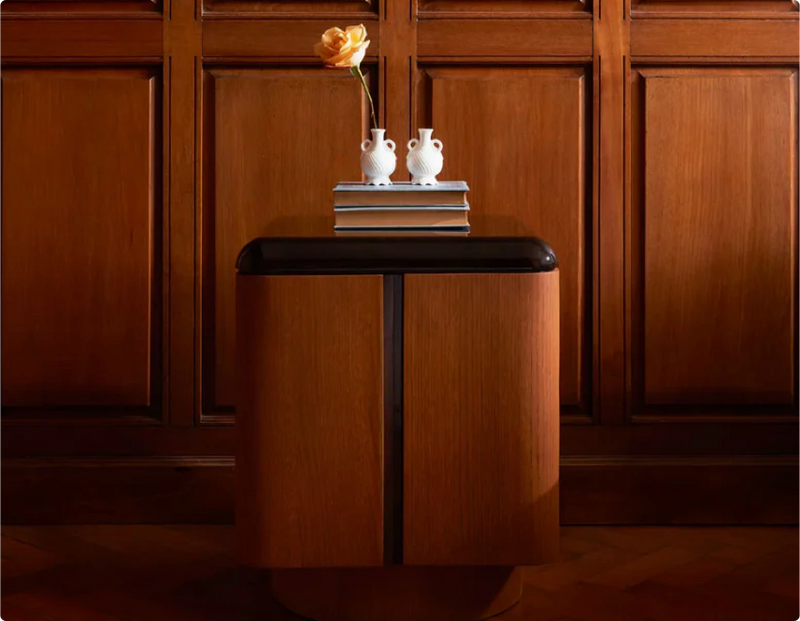 Stylish wooden side table with black accents, decorated with stacked books and two small white vases, placed against a wooden paneled wall.