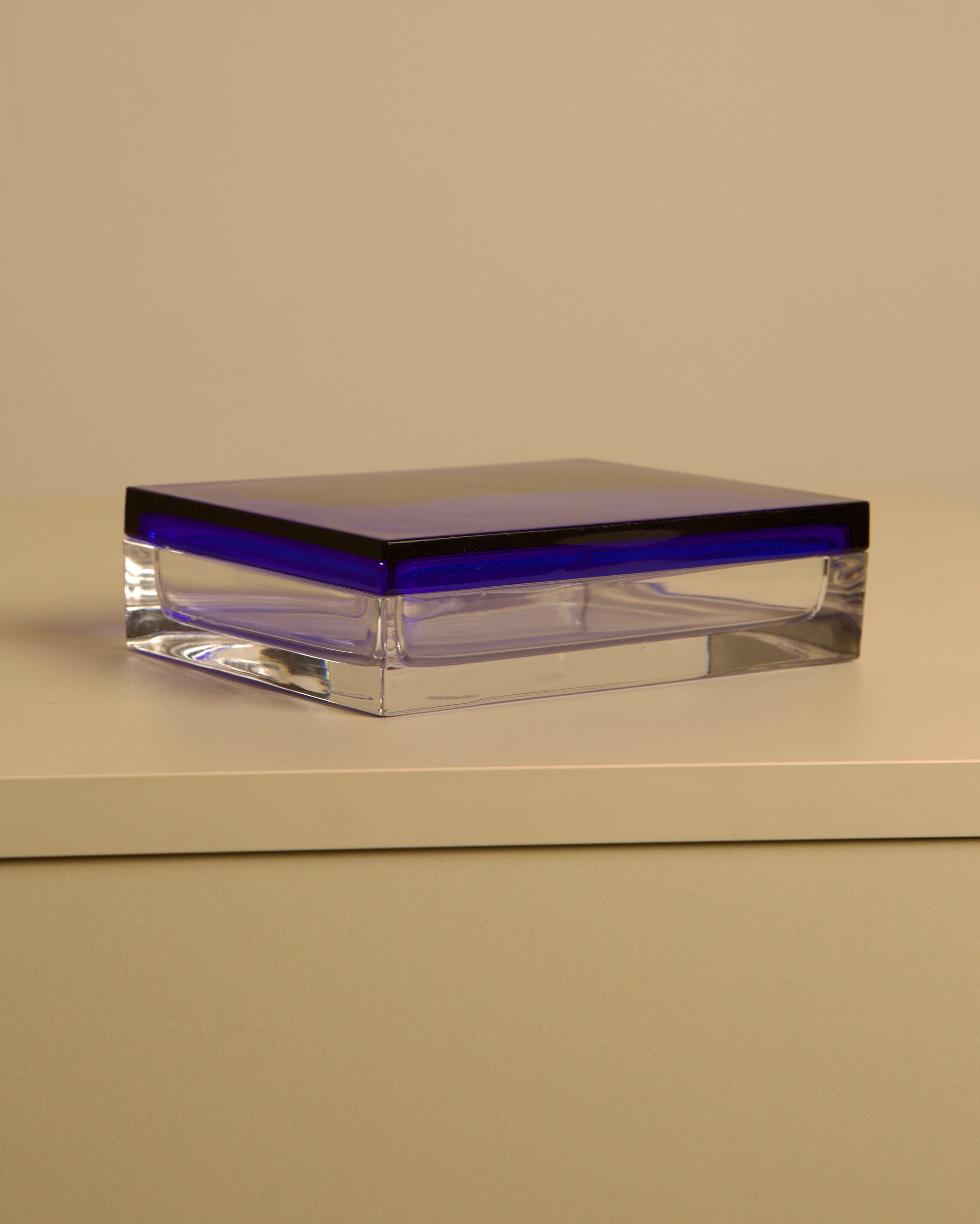 The Treaptyque Italian Box in transparent and blue crystal by Duccio Di Segna from the 80’s sits on a smooth, light surface with a plain beige background, showcasing its rectangular shape and striking deep blue lid.