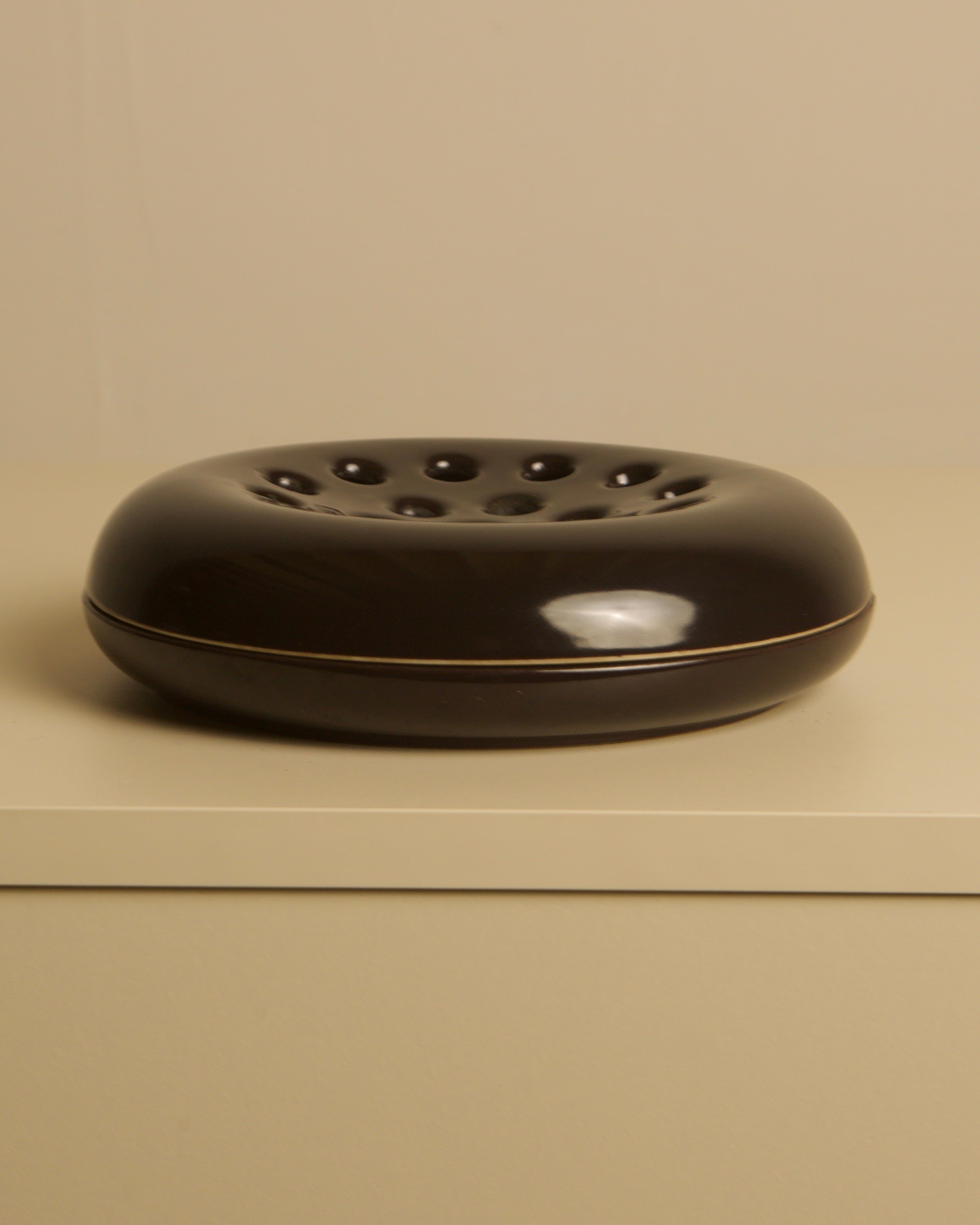 The Italian Brown Ceramic Ashtray by Angelo Mangiarotti for Brambilla (1960s, Treaptyque) features a round, dark brown glazed design with a central depression and holes, shown on a light surface against a beige background.