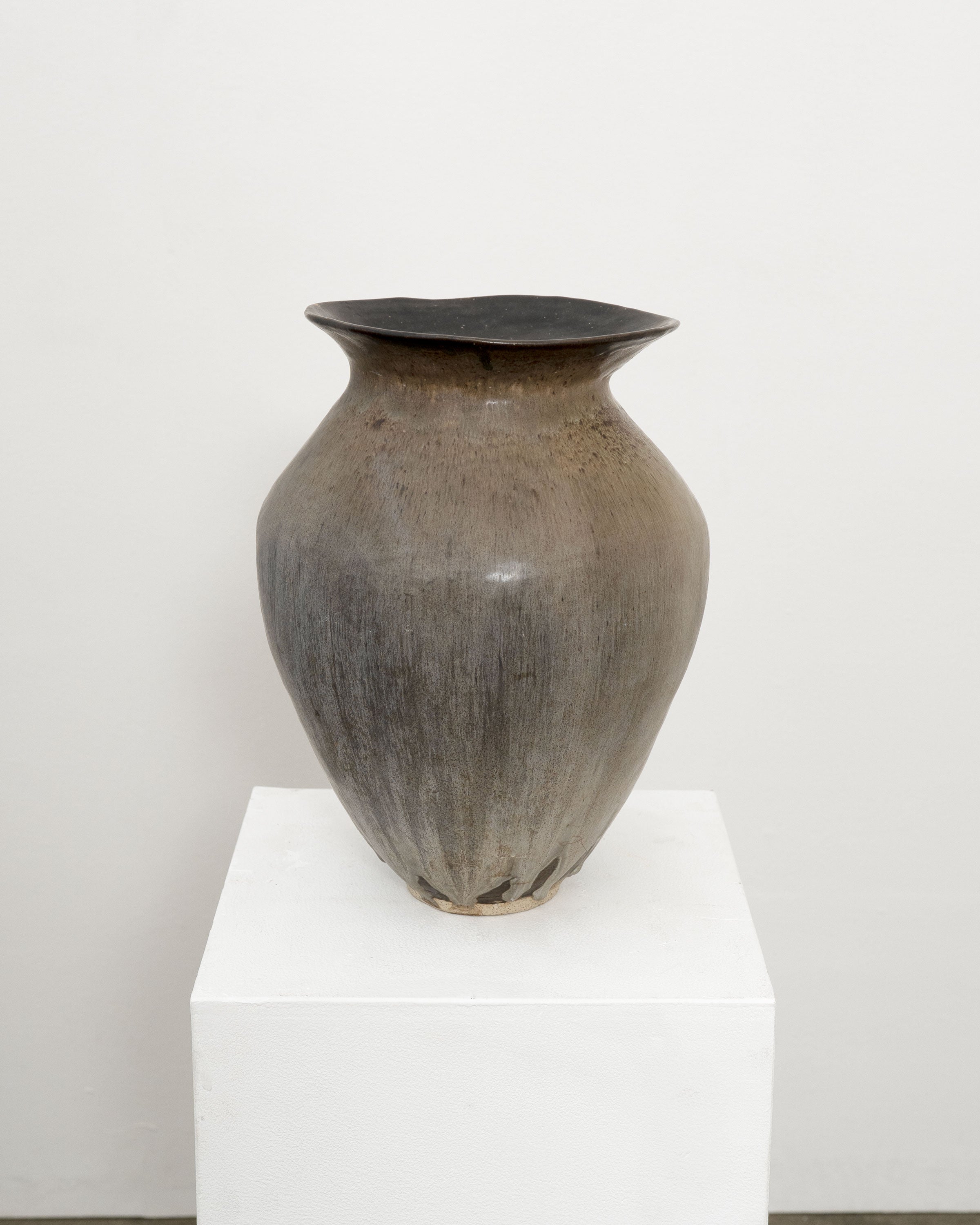 The Hand Built Studio Pottery Vase by Tom Bogle is a large, rounded brown stoneware vessel with a wide rim, narrow neck, and smooth matte finish. It sits on a white pedestal against a plain white background.