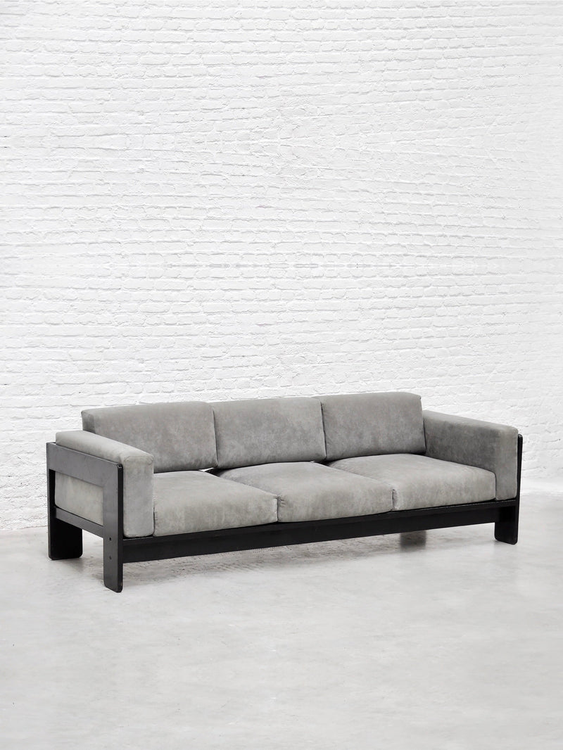 The Bastiano Sofa by Tobia Scarpa for Gavina, 1960s—available from Introverso—features gray suede cushions and a black wood frame, shown in a minimalist room with a white brick wall and concrete floor.