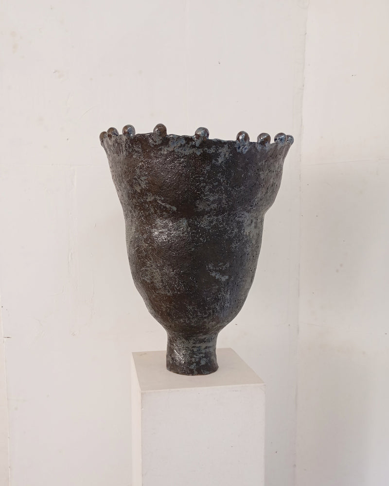 The Coupe Lilibet 7 by Claire Pain is a dark, textured ceramic vase with a narrow base, flared rim, rounded bumps along the top, and a subtle metallic glaze, shown on a white pedestal against a plain white wall.