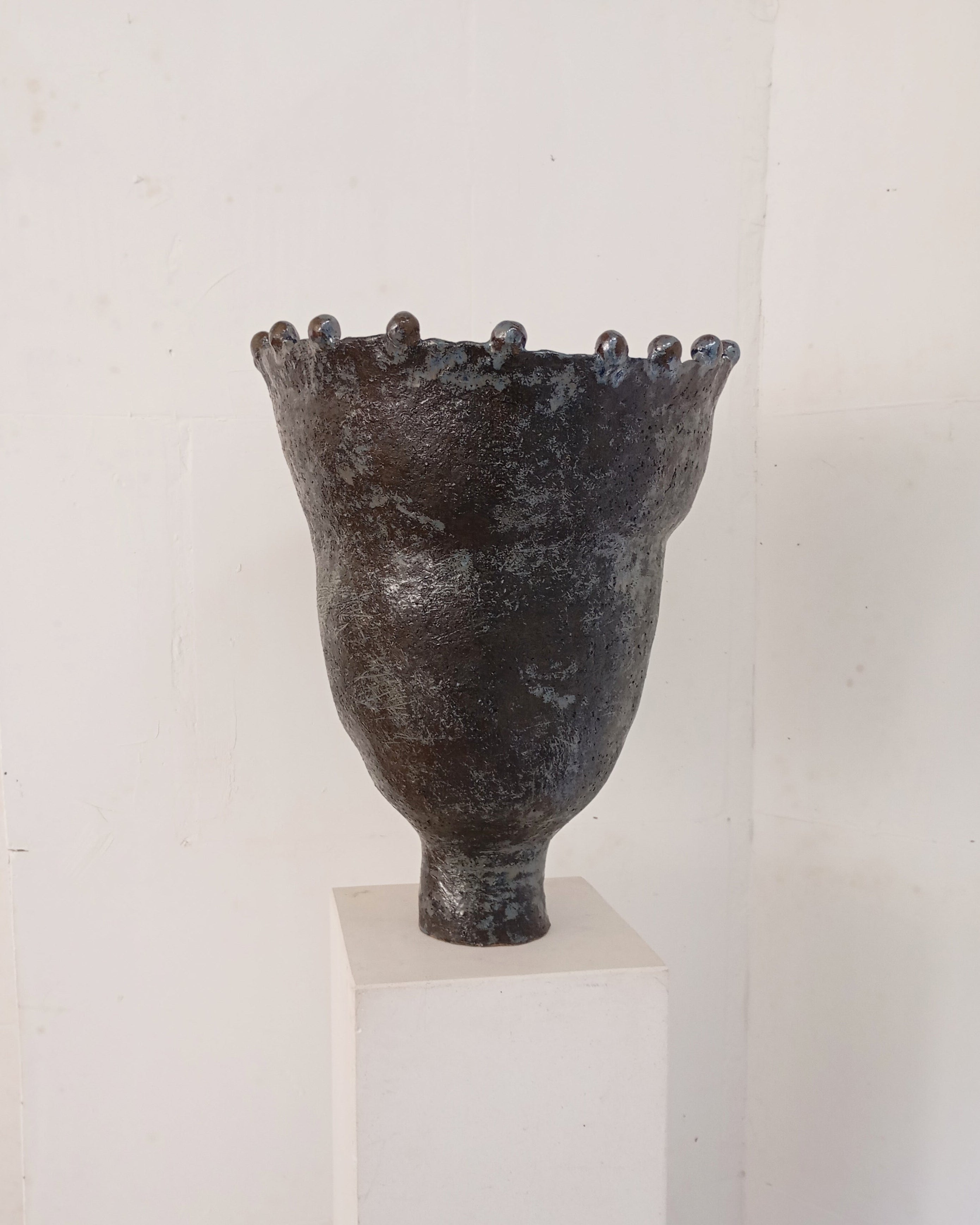 The Coupe Lilibet 7 by Claire Pain is a dark, textured ceramic vase with a narrow base, flared rim, rounded bumps along the top, and a subtle metallic glaze, shown on a white pedestal against a plain white wall.