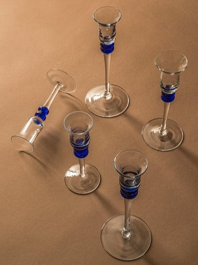 Five elegant Memphis Liquor Glasses with blue accents on the stems, arranged on a beige surface, with one glass tipped over, serving as unique candle holders. (Les Objets Amsterdam)