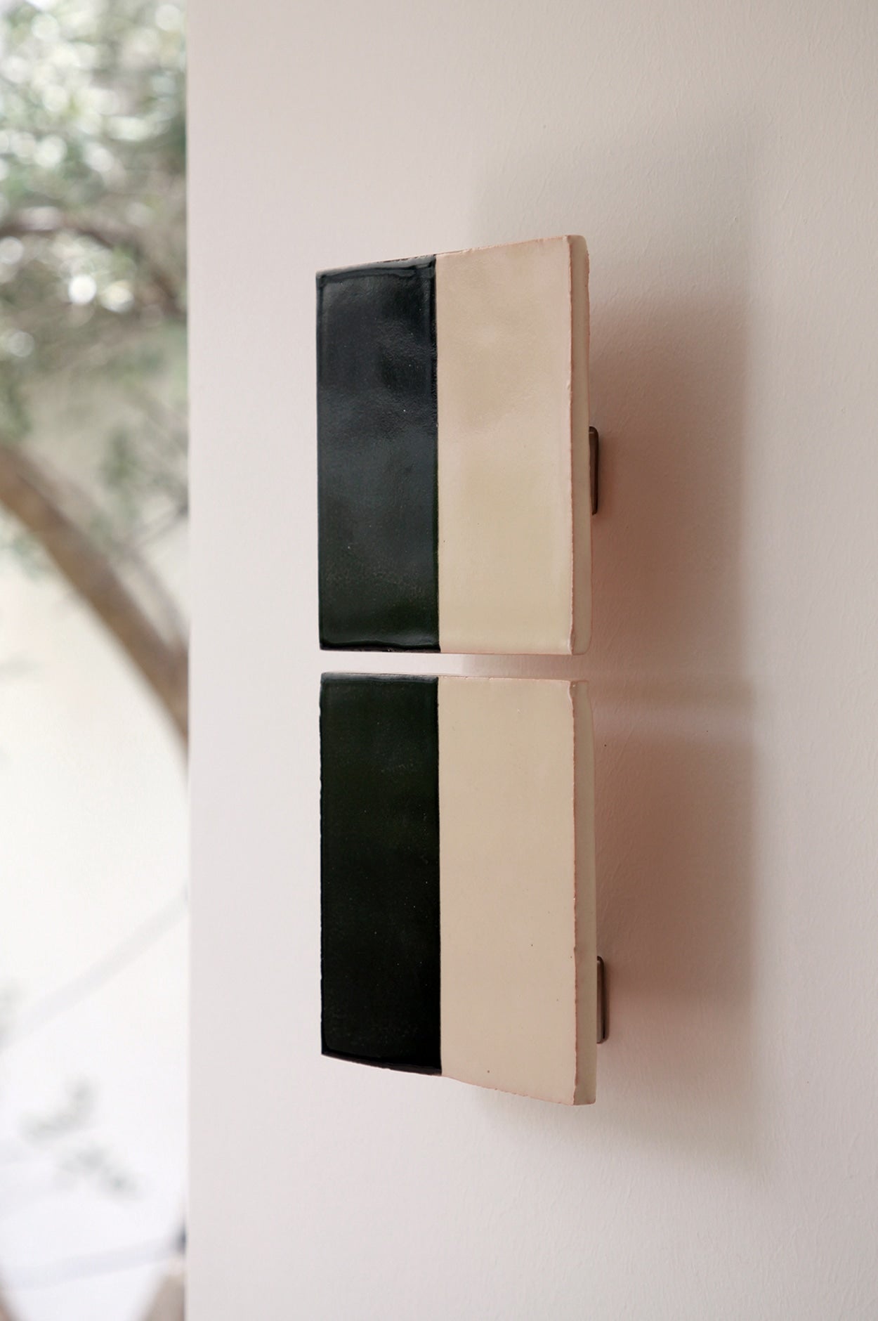 Two Tiles Line N wall appliques by Violaine d'Harcourt, each vertically split into black and cream, are mounted side by side on a white wall near a window with blurred tree branches outside—handmade design.