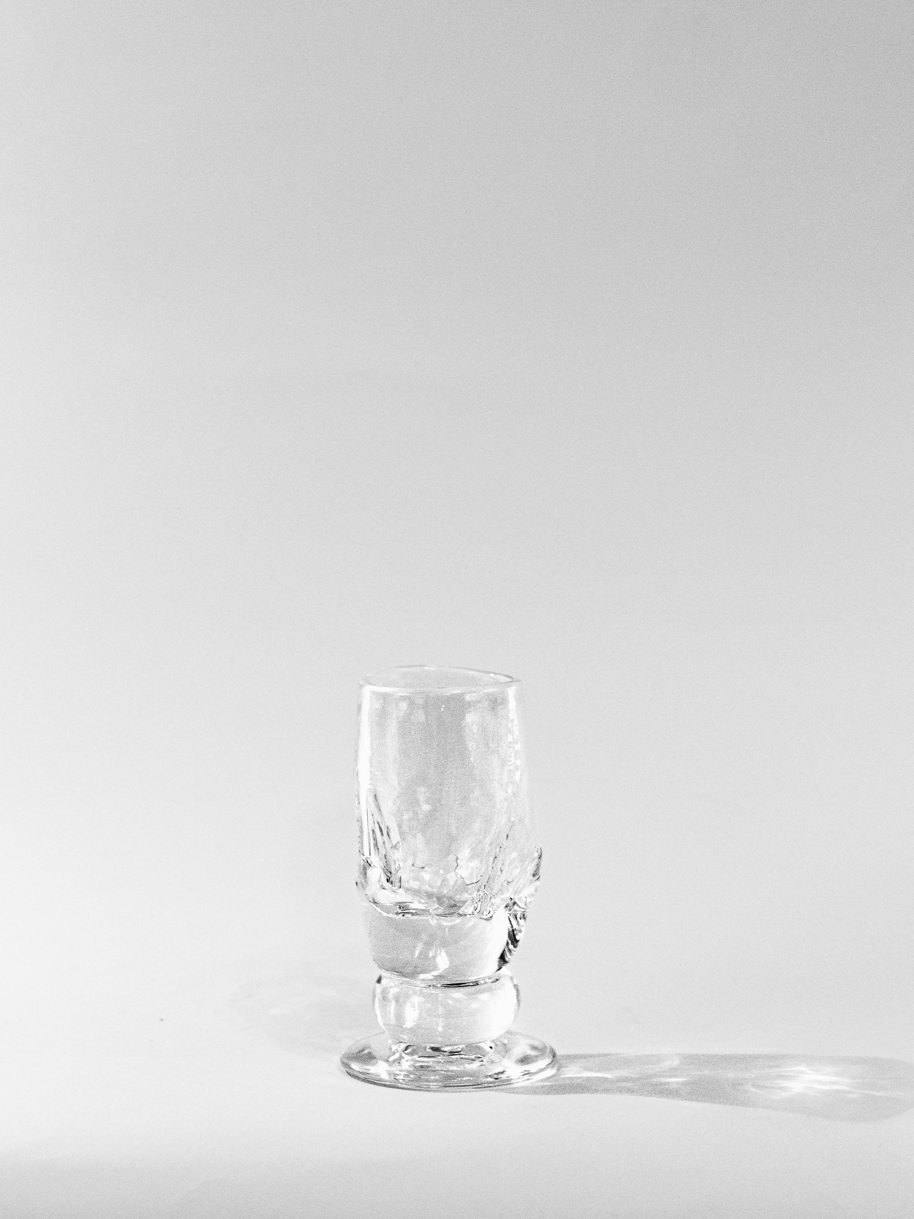 The Organic Footed Table Glass by Szkło Studio, featuring an irregular, textured hand-blown design, stands on a white surface against a plain light background, casting a soft shadow to the right.