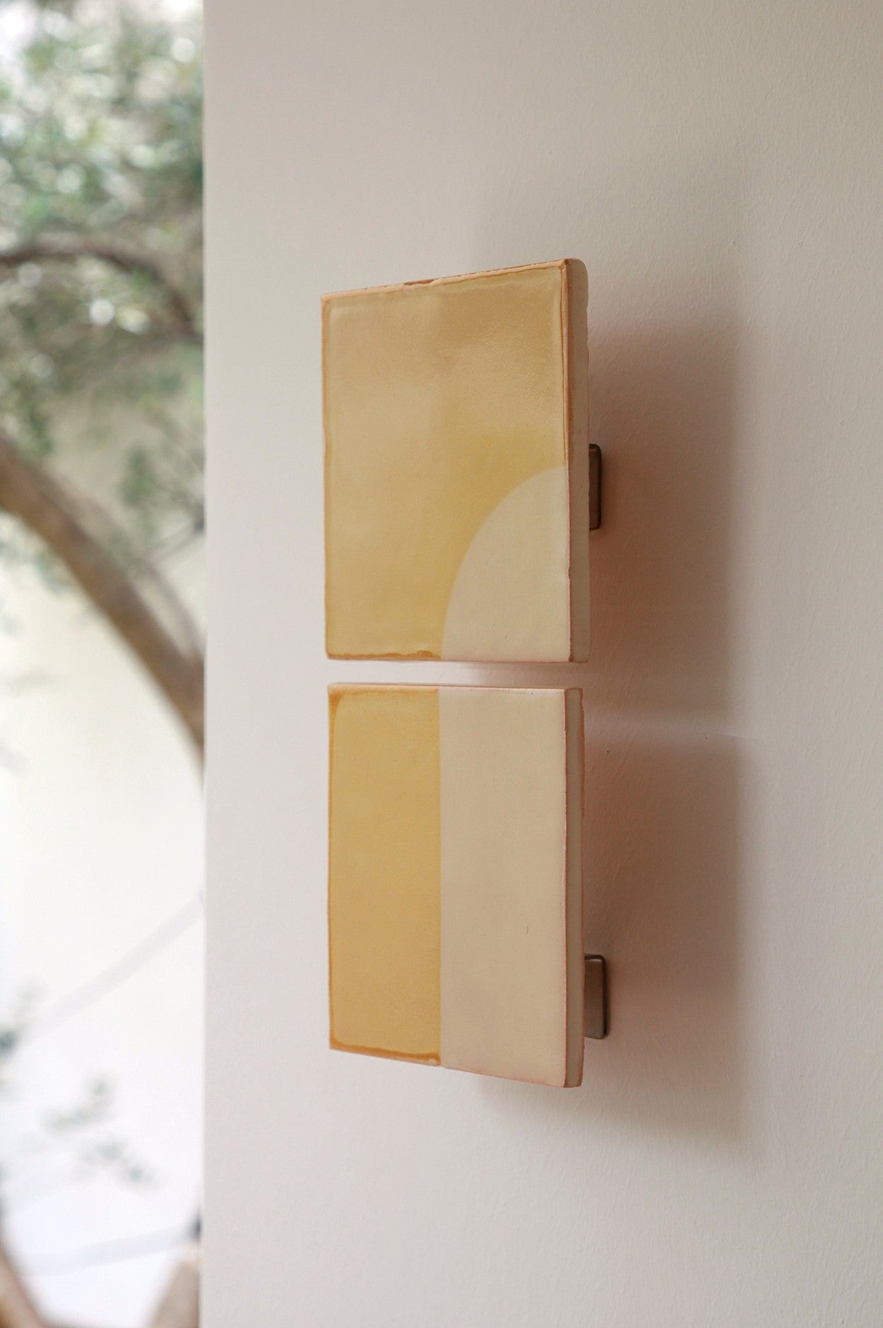 Two minimalist ceramic wall sconces from Violaine d'Harcourt evoke handcrafted tiles with soft yellow and cream geometric designs. Mounted on a white wall, they glow gently as blurred greenery filters through a Tiles Door J.