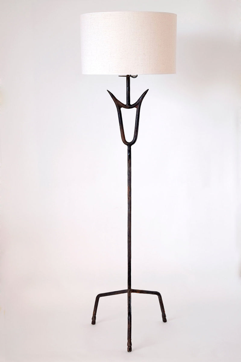 The "Sevilla" Lamp by Barracuda Interiors is a tall tripod floor lamp with a white drum shade, dark metal stand featuring a forked design near the top, and three angled legs, shown against a plain white background.