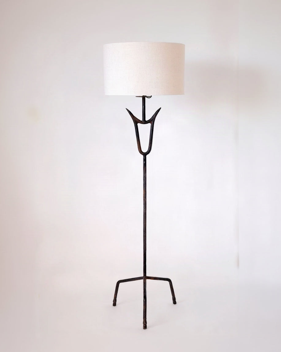 The "Sevilla" Lamp by Barracuda Interiors is a tall tripod floor lamp with a white drum shade, dark metal stand featuring a forked design near the top, and three angled legs, shown against a plain white background.