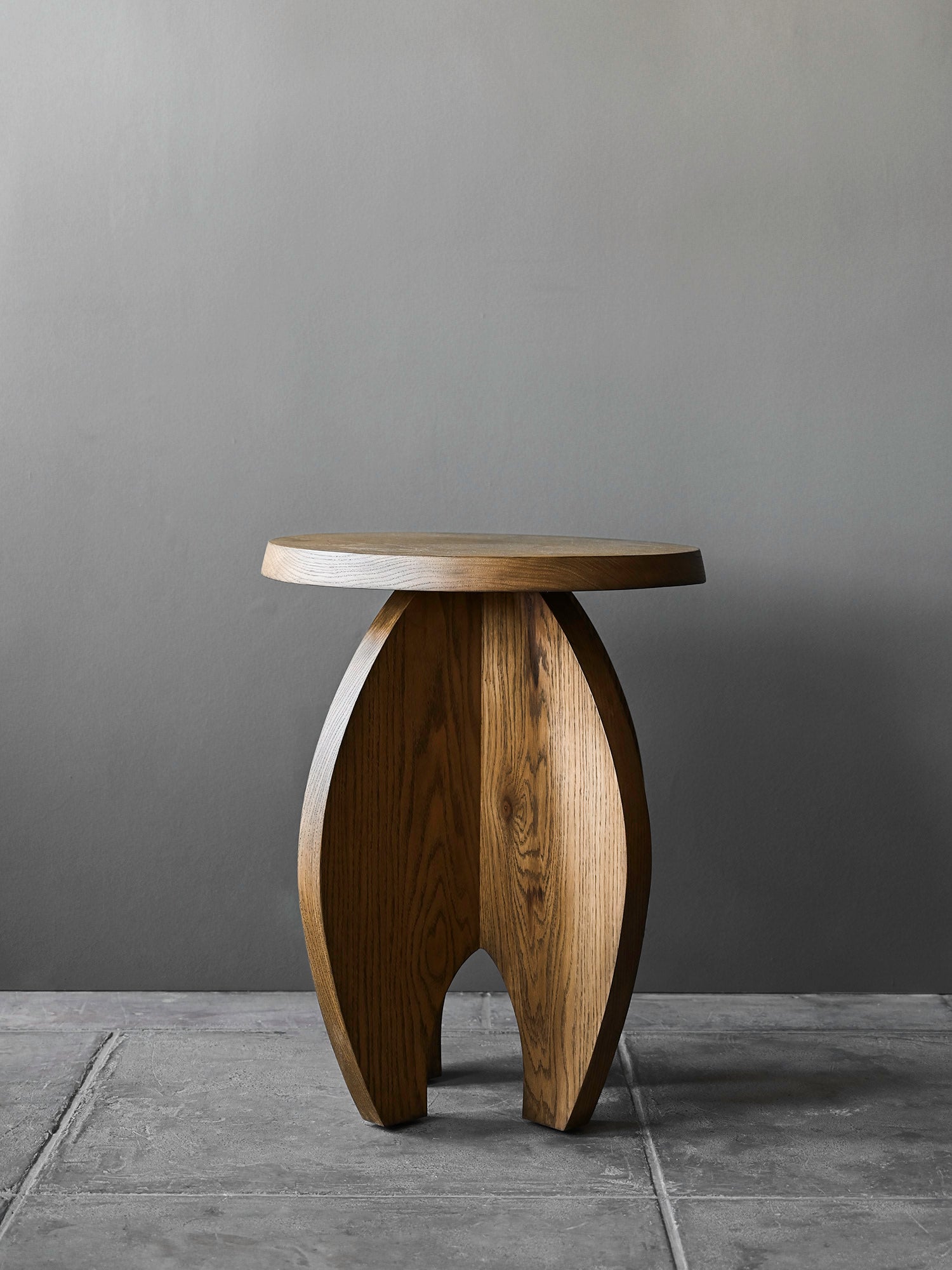 The 'Fusée' Stained Oak Side Table by Studio OSKLO features a modern, round thick top and two interlocking rocket-shaped legs, displayed against a gray wall and stone floor.