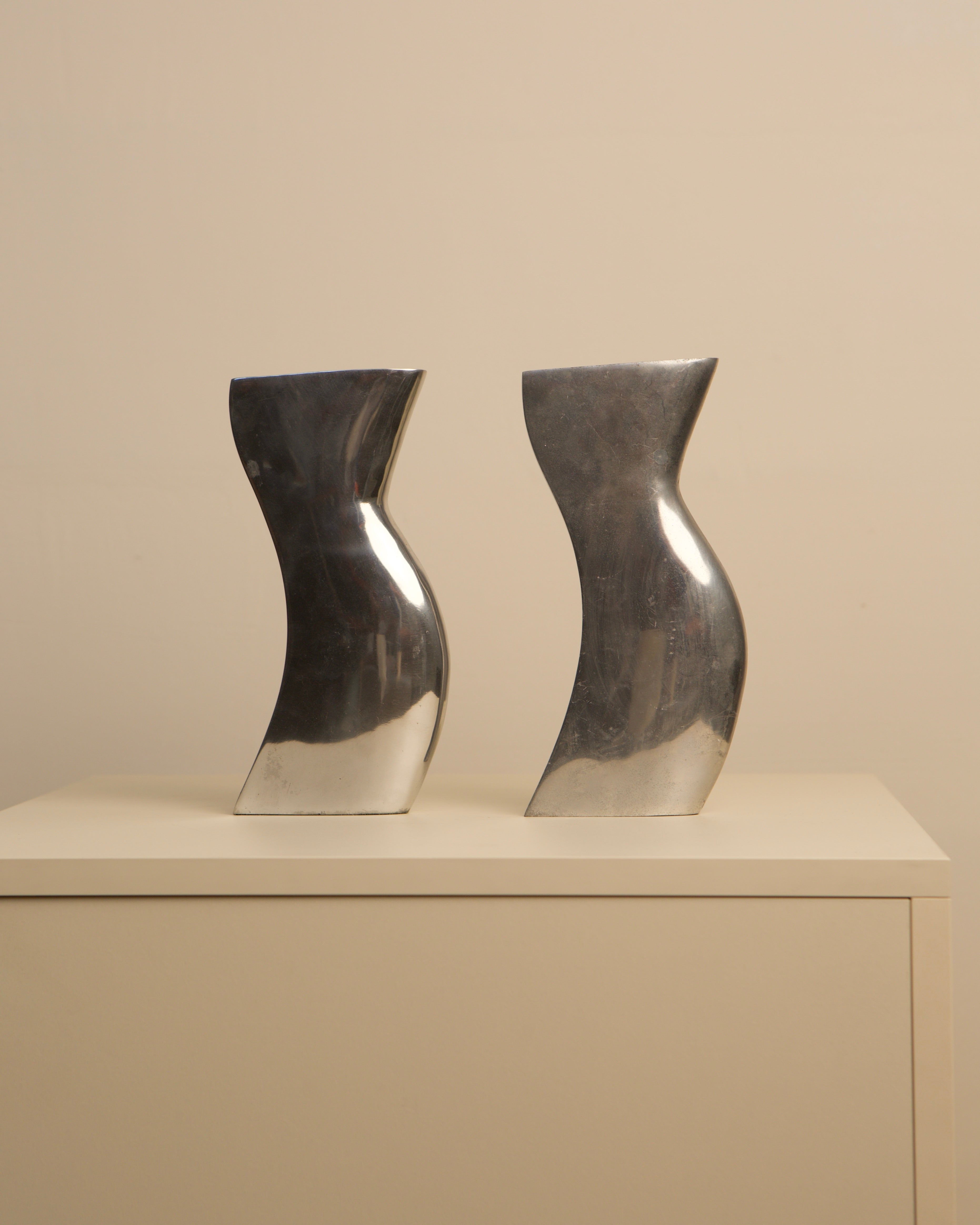 Pair of curved aluminum Dance vases on beige surface, side view