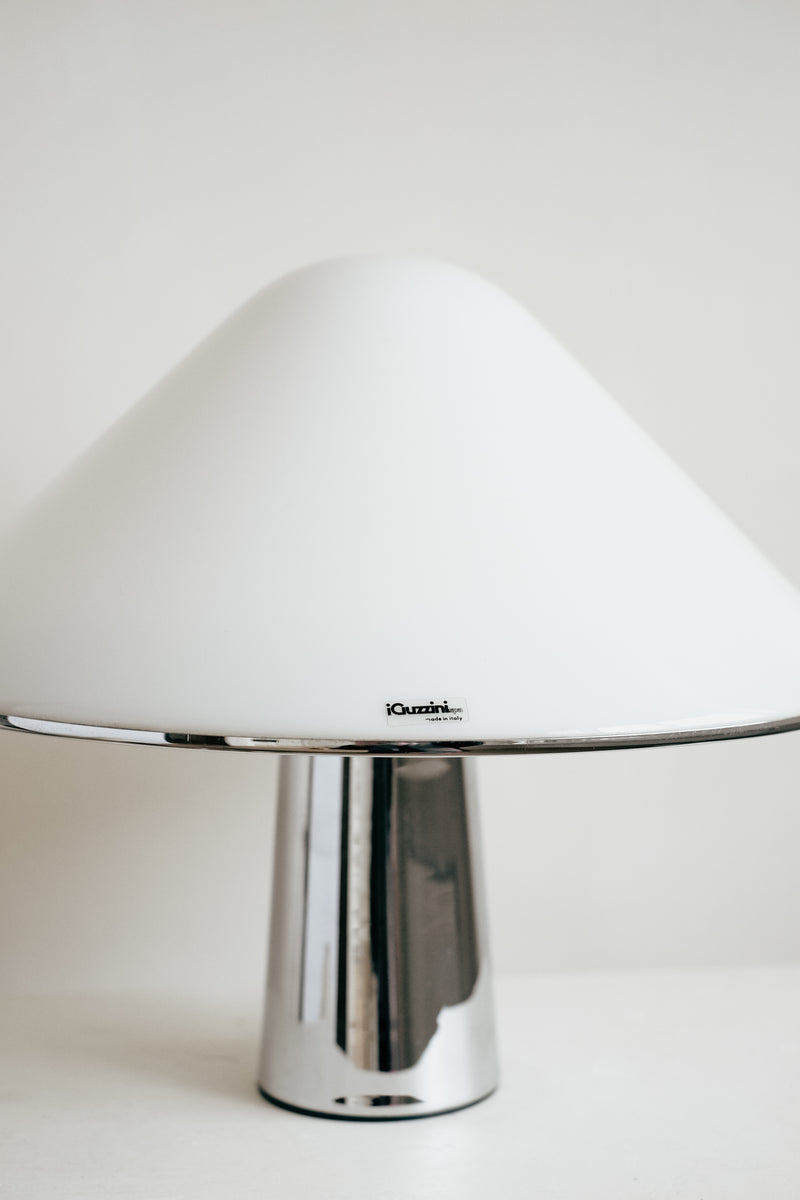 The Harvey Guzzini Elpis Table Lamp by septembre studios features a wide white dome-shaped shade and a shiny cylindrical metal base, inspired by Luigi Massoni’s design, set against a plain light background.