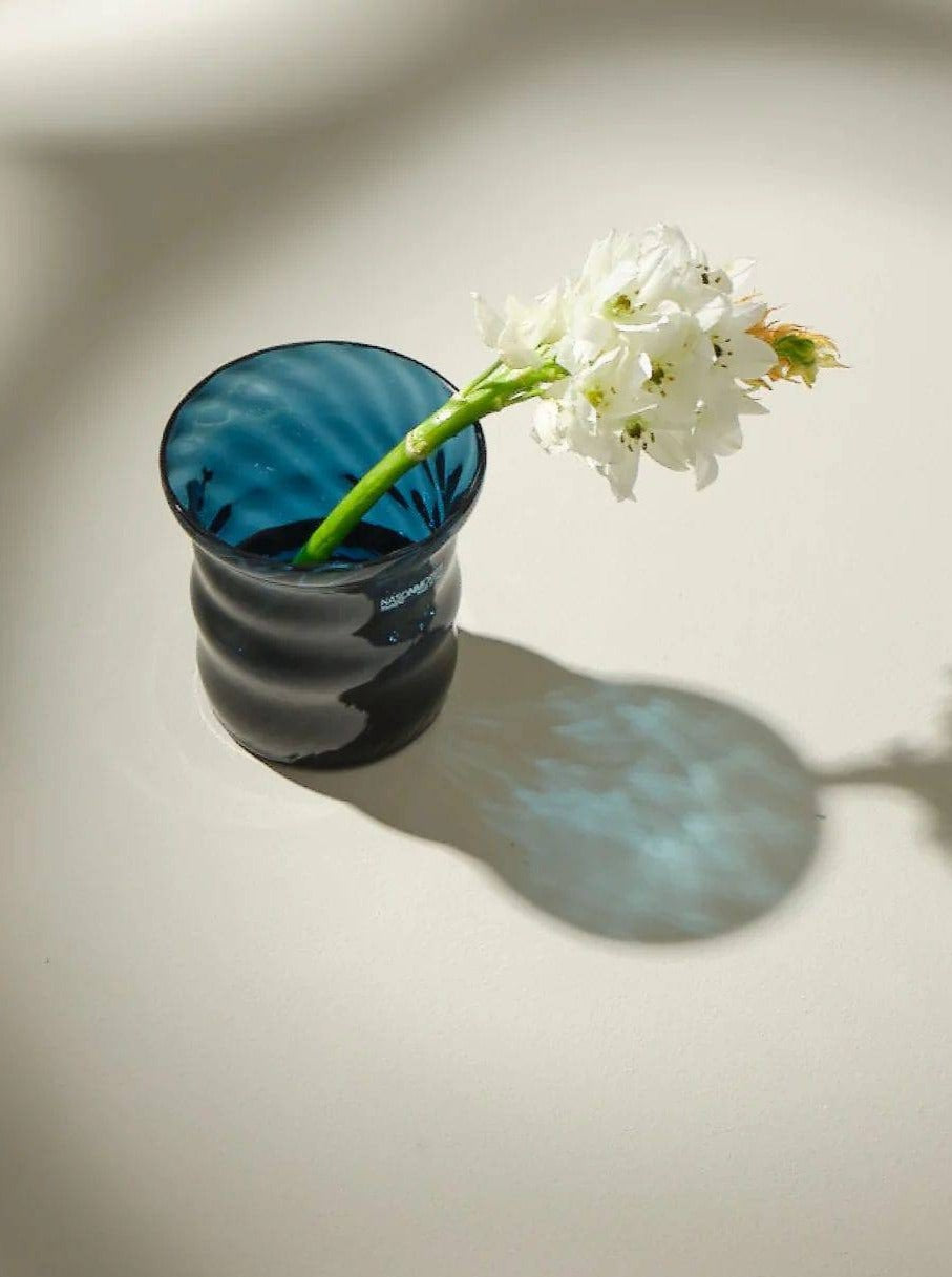 A single white flower with a green stem resting in a blue translucent glass vase, casting a soft wavy shadow on a light surface illuminated by sunlight. The vase features an intricate Venetian decorative motif holding The First Candle by Aina Kari.