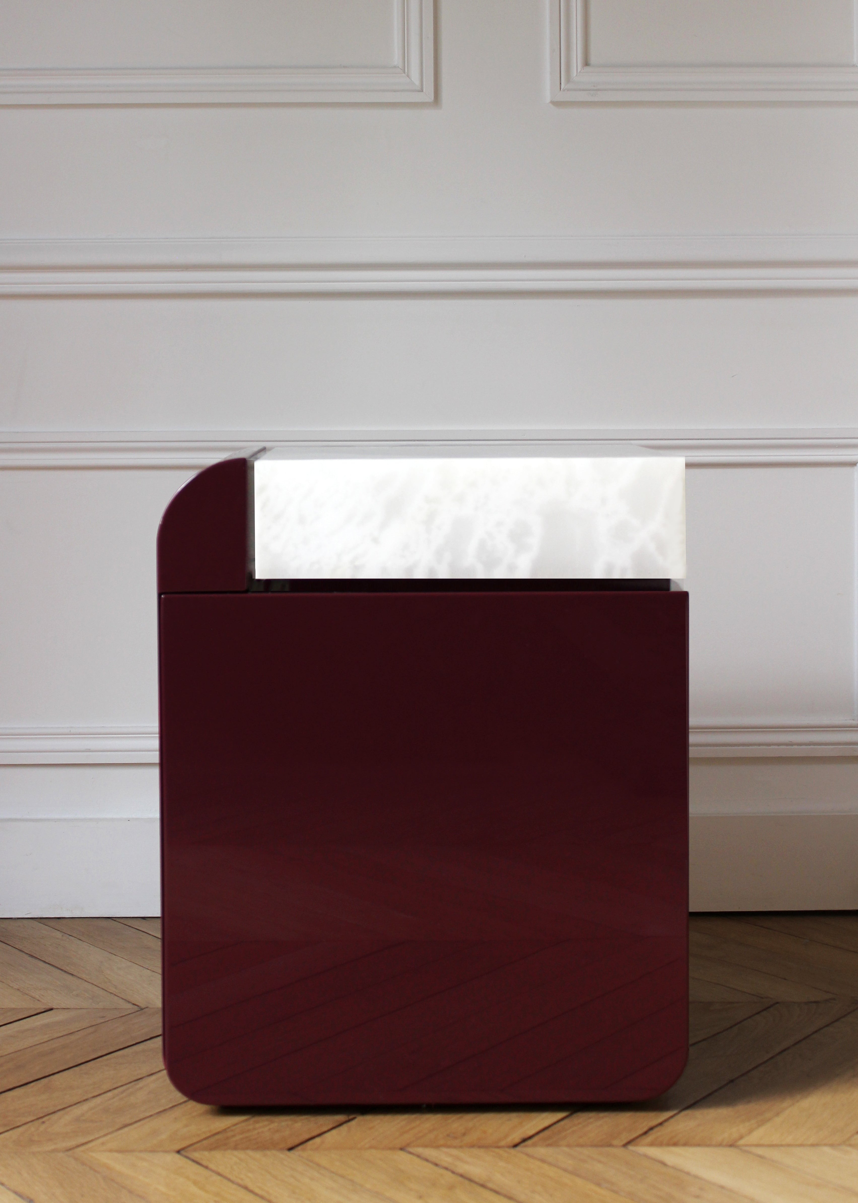 The "Amo" Bordeaux and White Onyx Table by Marbera rests beneath a modern burgundy lamp with a white illuminated top, set against a white paneled wall. This setup enhances the contemporary design of the herringbone-patterned wooden floor.