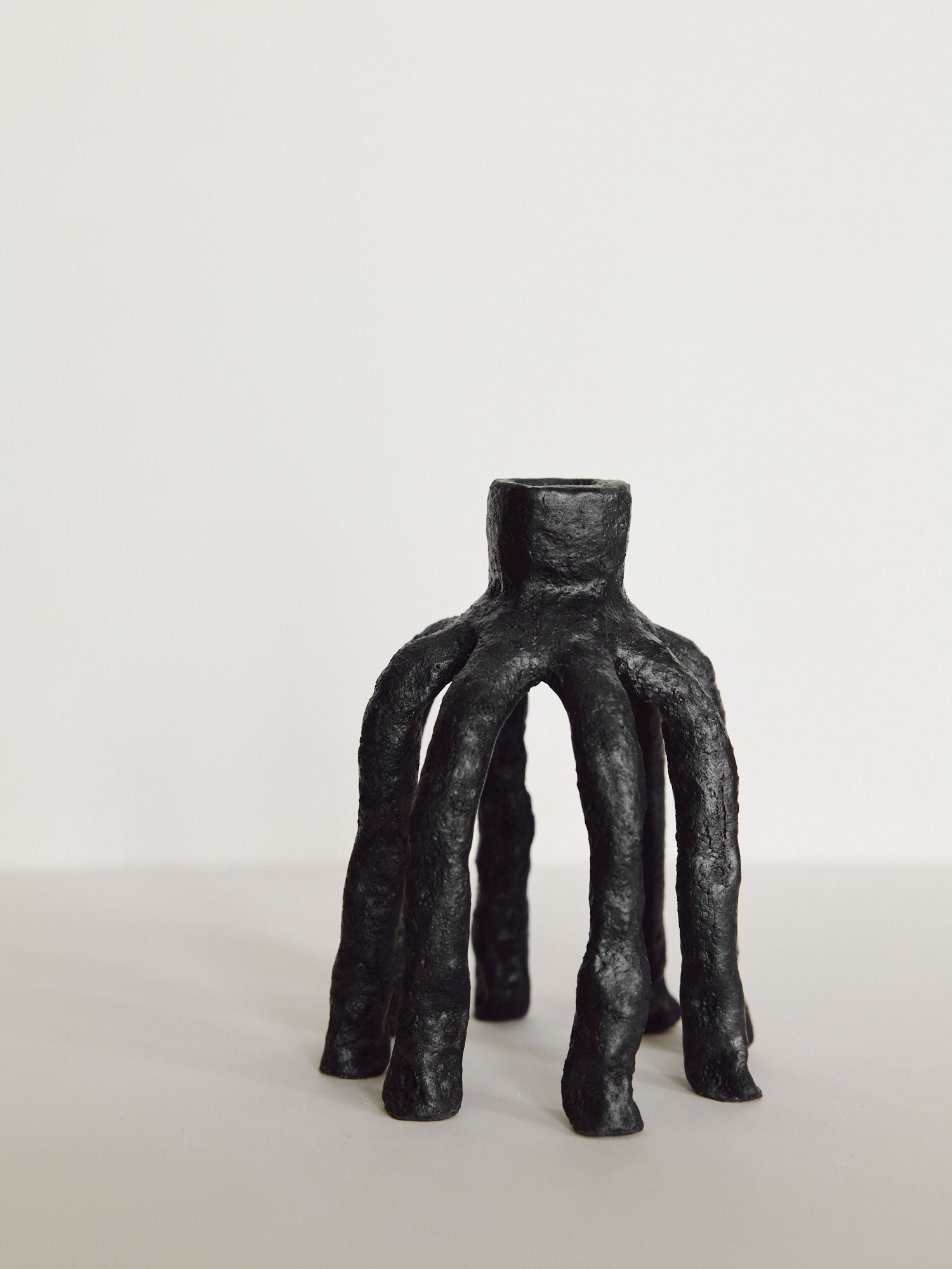 The Candleholder La Chose 1 by Claire Pain is a stoneware piece with five textured, organic legs, standing upright and finished in satin black glaze, displayed on a light surface against a plain background.