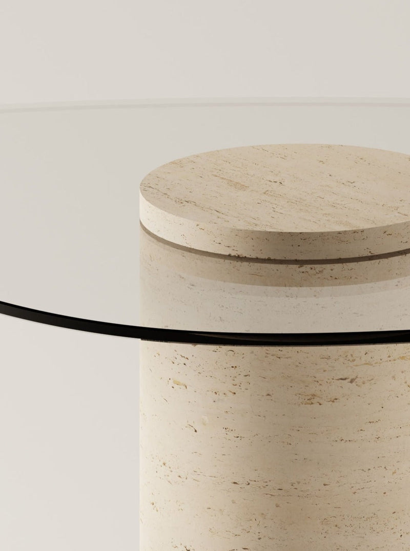 The UUfo 02 Dining Table by BURGIO. features a round glass top on a cylindrical beige stone base. Its modern, organic silhouette and minimalist appeal are highlighted in a close-up against a plain, light background.