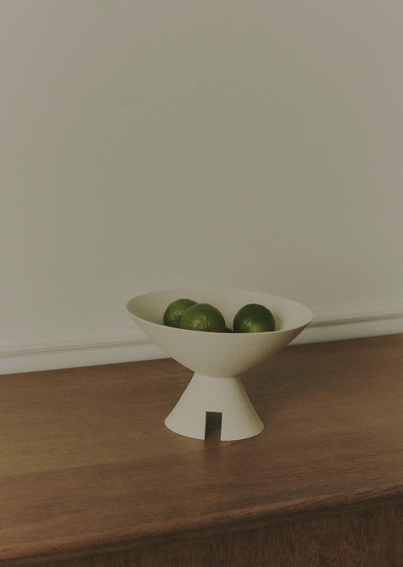 A minimalist image of a white, Argot Studio Budō Bowl on a wooden surface, containing several green limes. The background is a plain, light-colored wall.