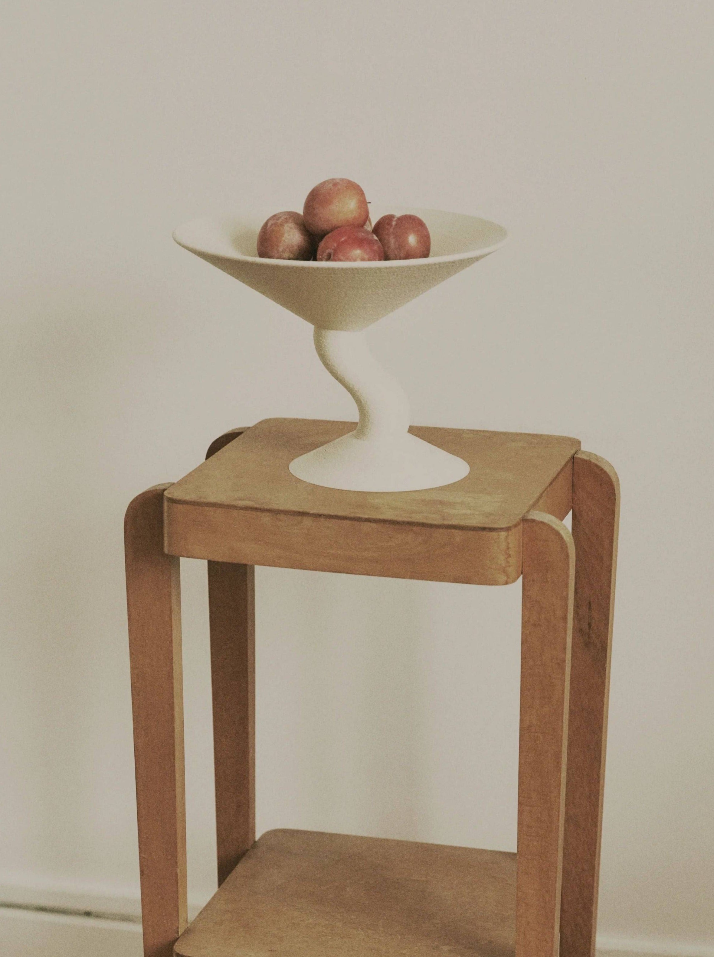 A white pedestal decorative bowl containing apples on a Argot Studio Wavy Fruit Stand against a plain wall.