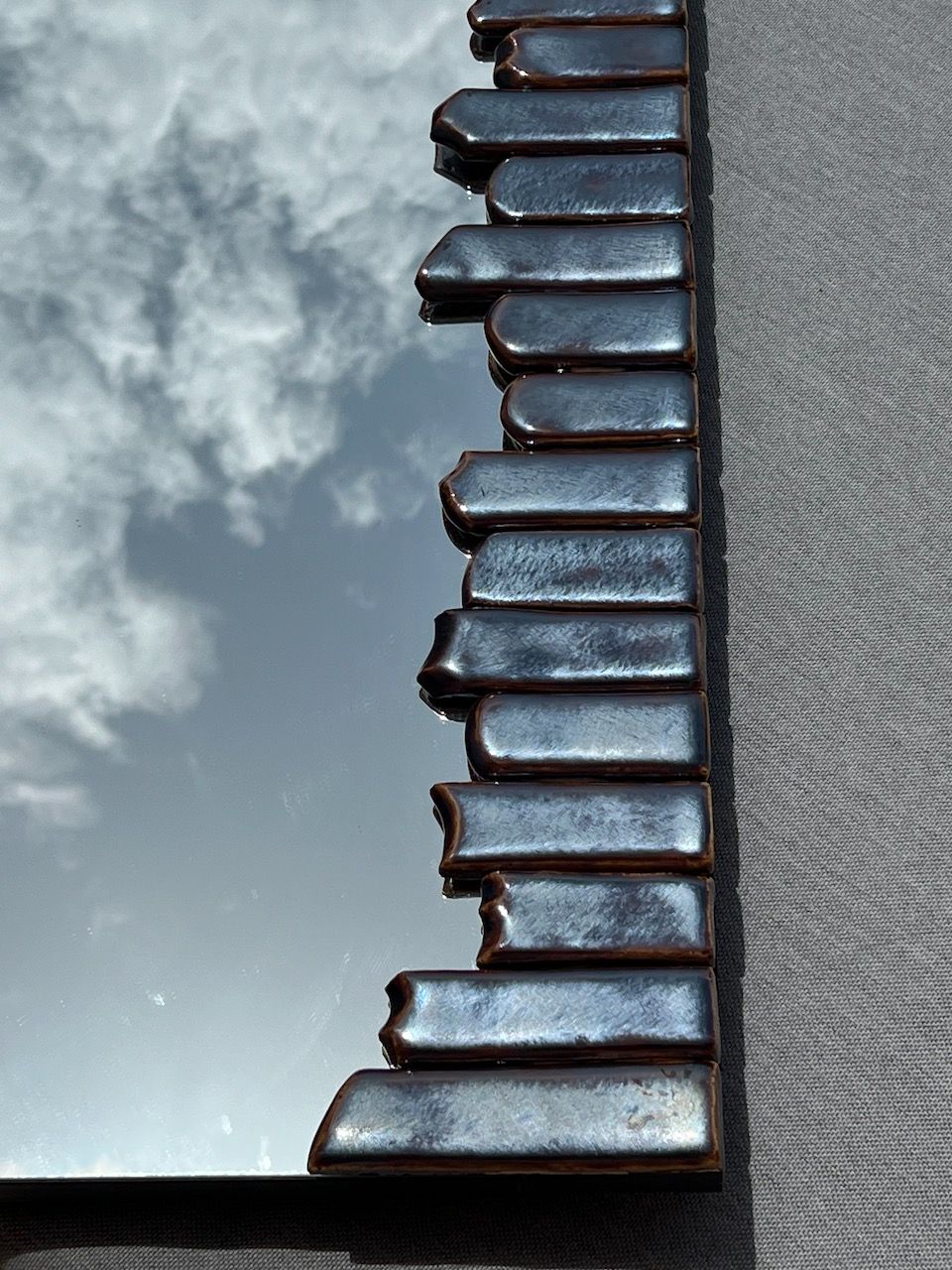 A close-up of a Katia Langeard Cliff Collection Contemporary Mirror in ceramic, featuring a sculpted dark frame that reflects a partly cloudy sky against a textured gray background.