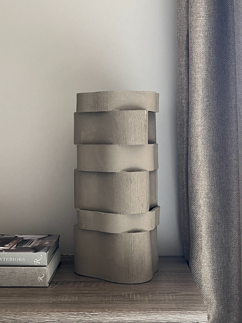 A large, modern textured gray vase with wavy patterns sits on a wooden surface beside a stack of books, a Katia Langeard 'Too 60' Ceramic Table Lamp, and a gray curtain.