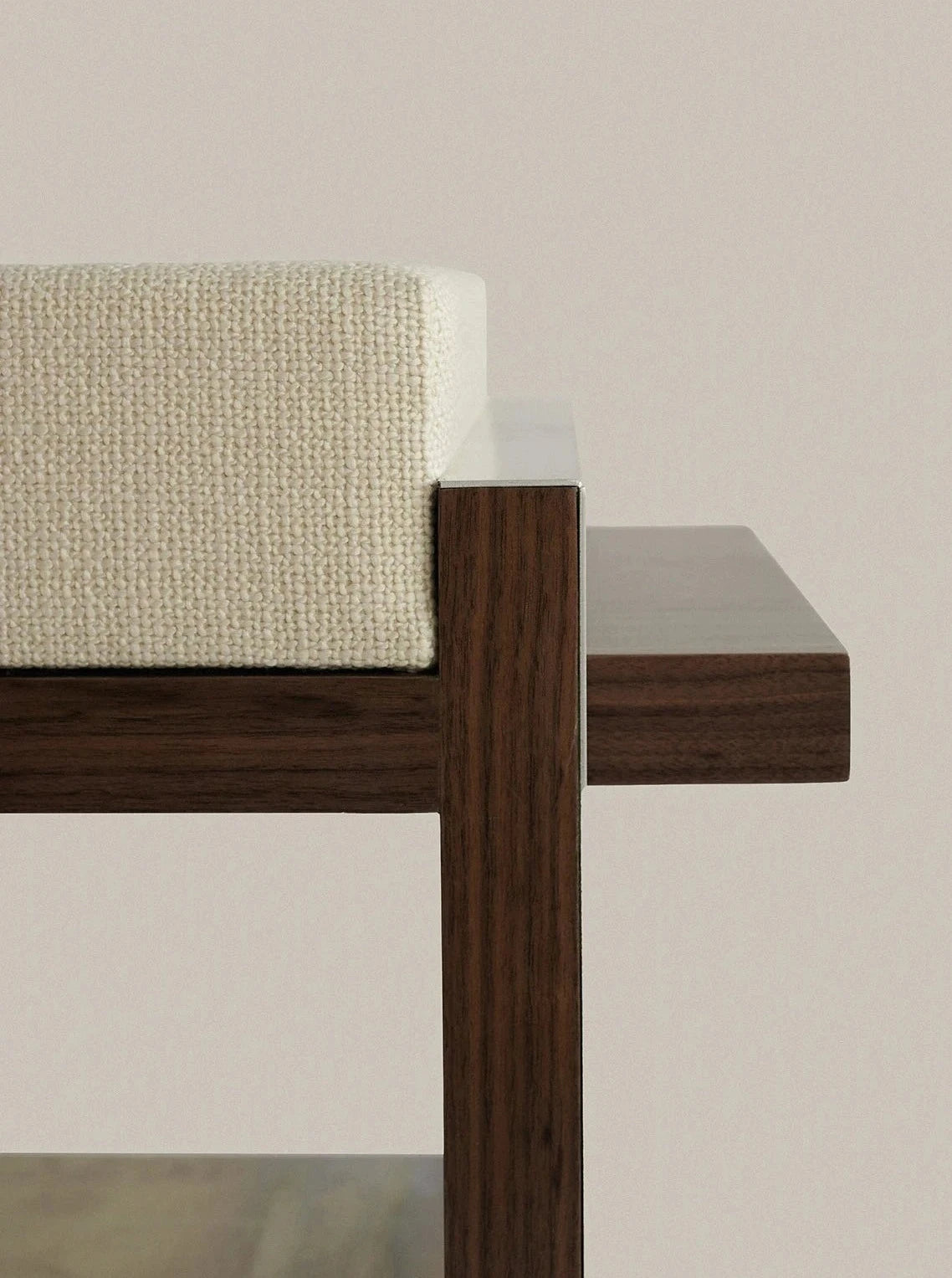 A close-up view of a Sofia De=Francesco B1 [single stool] with a light-colored cushioned seat. Crafted from walnut wood, the design showcases clean, minimalist lines. The dark finish contrasts beautifully with the textured fabric cushion, highlighting its artisanal appeal.