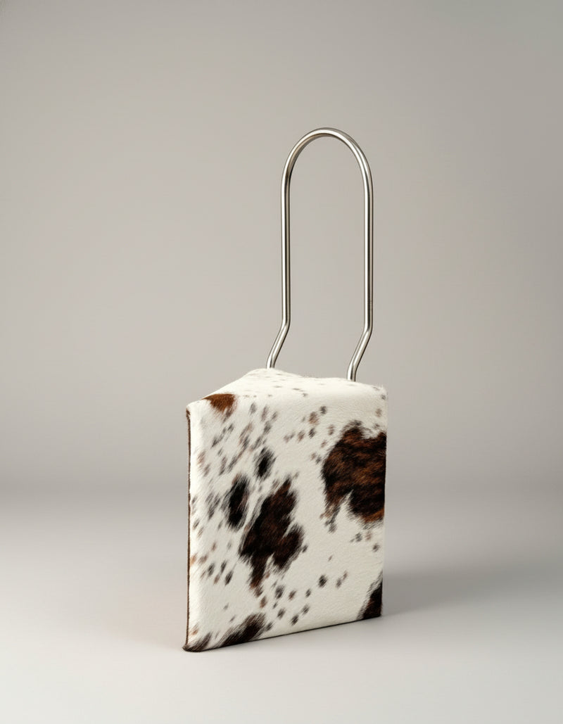The "Twist" Chair by Animate Objects features a rectangular body in white faux fur with brown and black cow print spots, reminiscent of cowhide upholstery, and is accented by a tall, thin silver metal handle. The background is plain and neutral.