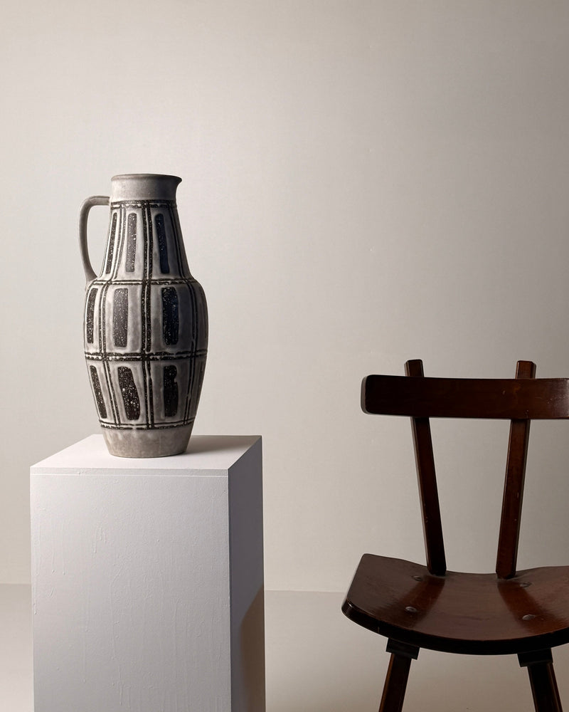 Ceramic pitcher, Germany, circa 1960. — angled view on white pedestal with wooden chair