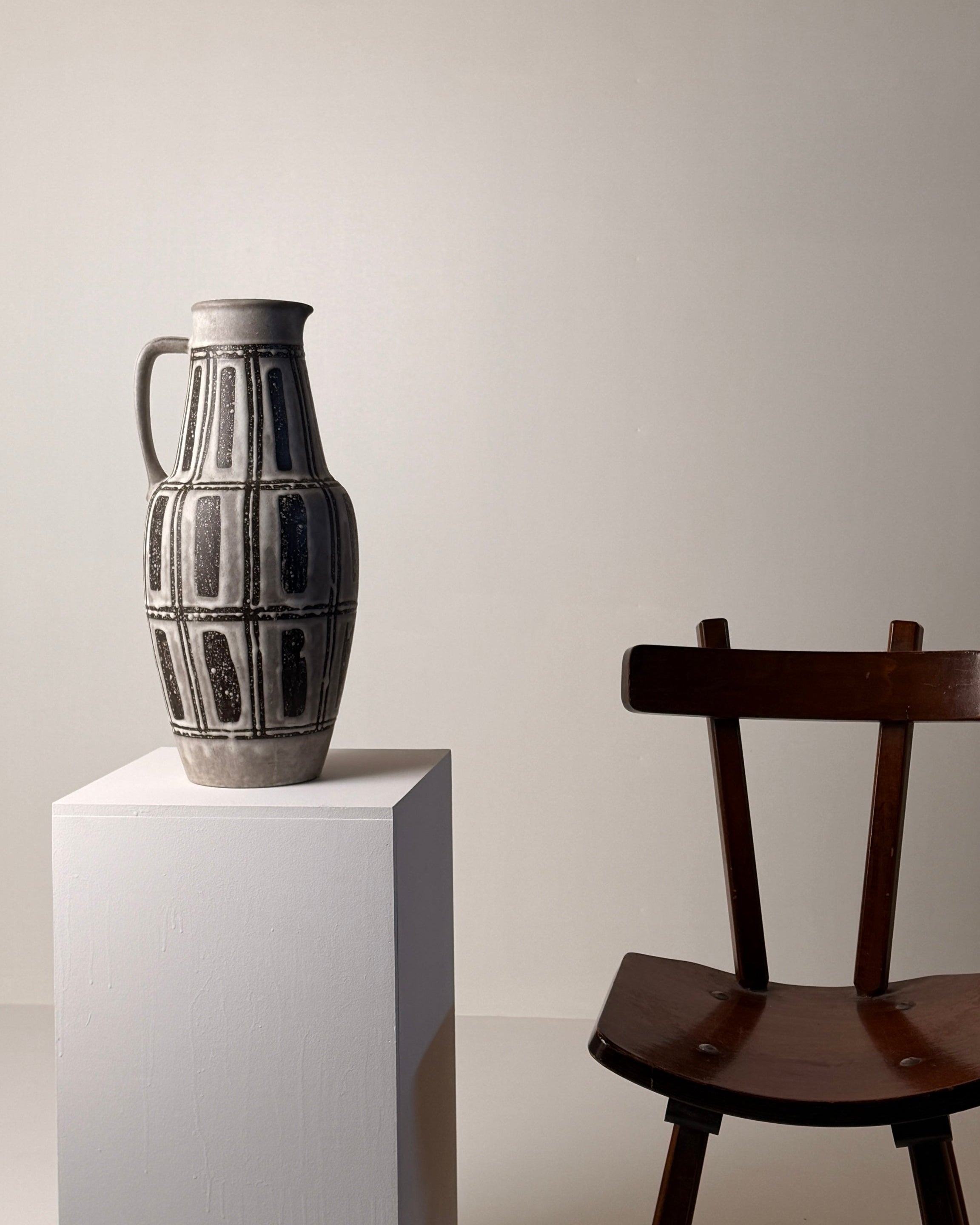 Ceramic pitcher, Germany, circa 1960. — angled view on white pedestal with wooden chair