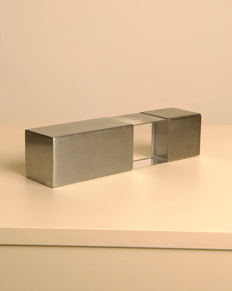 The Treaptyque Italian Sculpture in Metal and Plexiglass 70's features brushed metal ends and a transparent plexiglass center, displayed on a light surface with a beige background, reflecting the sleek elegance of 1970s Italian design.